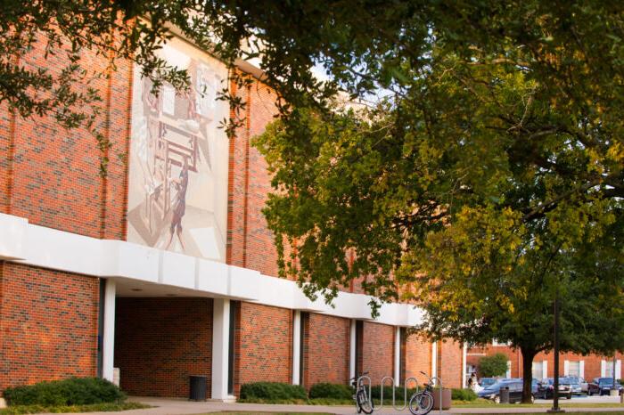 Brick building with large mural visible, framed by leafy green trees. White architectural details contrast with red bricks. Sunlight casts warm glow on facade. Campus or institutional setting.