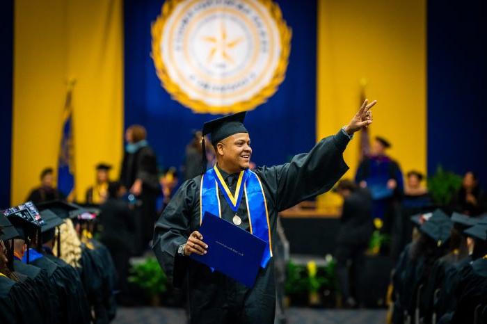 A male graduate in a cap and gown smiles and waves while holding his diploma.